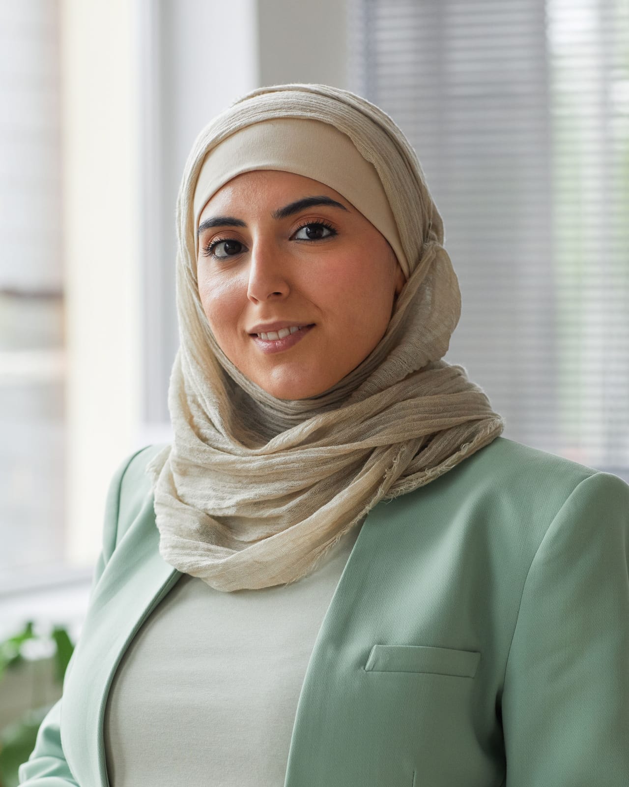 امرأة مبتسمة ترتدي حجابًا وسترة خضراء فاتحة تقف في مكتب مشرق